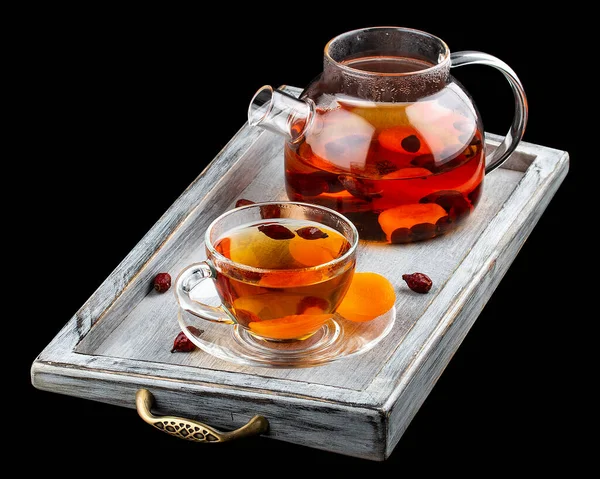Hot tea with rose hips and dried apricots in a glass teapot. On dark background