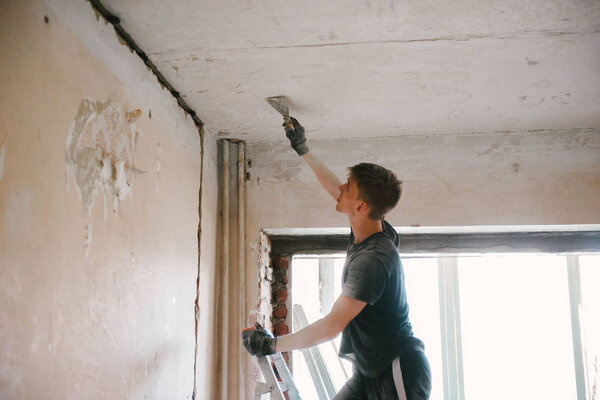 A man removes old paint from the ceiling with a spatula