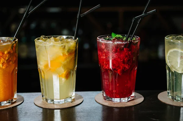 Verschillende soorten zomerdrankjes in glazen, ijsblokjes en schijfjes fruit op een donkere, brede tafel. — Stockfoto