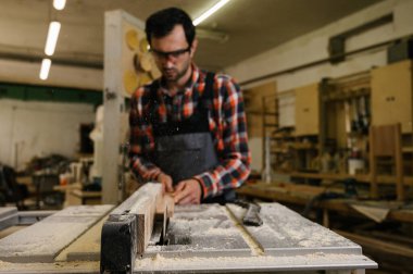 Marangozluk atölyesinde çalışma süreci. Marangozluk atölyesinde çalışan bir adam. Marangoz, kereste fabrikasında tahta kesmek için testere makinesi kullanıyor. Meslek, marangozluk, marangozluk, marangozluk.