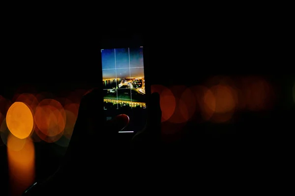 A man takes a photo of the night city on a smartphone.Shooting the city lights with a mobile device at night. View through the screen at the time a man takes a photo of a city at night. Cityscape