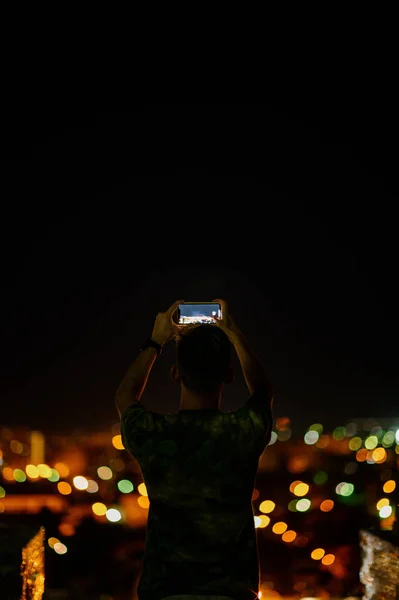 A man takes a photo of the night city on a smartphone.Shooting the city lights with a mobile device at night. View through the screen at the time a man takes a photo of a city at night. Cityscape