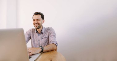 Tişörtlü, elinde kahve olan mutlu adam dizüstü bilgisayarla çalışıyor. Genç adam evde, masada dizüstü bilgisayarla çalışıyor. Genç adam not defteri üzerinde çalışıyor. Dizüstü bilgisayarla proje üzerinde çalışıyor.