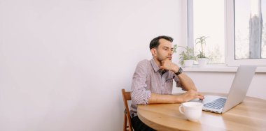 Gömlekli ve kahveli bir adam dizüstü bilgisayarla çalışıyor. Genç adam evde, masada dizüstü bilgisayarla çalışıyor. Genç adam not defteri üzerinde çalışıyor. Dizüstü bilgisayarla proje üzerinde çalışıyor.