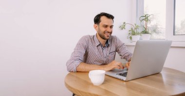 Tişörtlü, elinde kahve olan mutlu adam dizüstü bilgisayarla çalışıyor. Genç adam evde, masada dizüstü bilgisayarla çalışıyor. Genç adam not defteri üzerinde çalışıyor. Dizüstü bilgisayarla proje üzerinde çalışıyor.