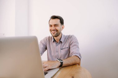 Tişörtlü, elinde kahve olan mutlu adam dizüstü bilgisayarla çalışıyor. Genç adam evde, masada dizüstü bilgisayarla çalışıyor. Genç adam not defteri üzerinde çalışıyor. Dizüstü bilgisayarla proje üzerinde çalışıyor.