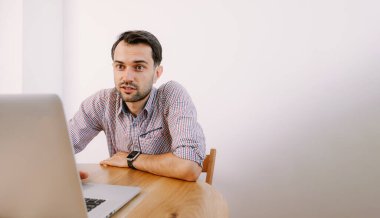 Gömlekli ve kahveli bir adam dizüstü bilgisayarla çalışıyor. Genç adam evde, masada dizüstü bilgisayarla çalışıyor. Genç adam not defteri üzerinde çalışıyor. Dizüstü bilgisayarla proje üzerinde çalışıyor.