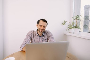 Tişörtlü, elinde kahve olan mutlu adam dizüstü bilgisayarla çalışıyor. Genç adam evde, masada dizüstü bilgisayarla çalışıyor. Genç adam not defteri üzerinde çalışıyor. Dizüstü bilgisayarla proje üzerinde çalışıyor.