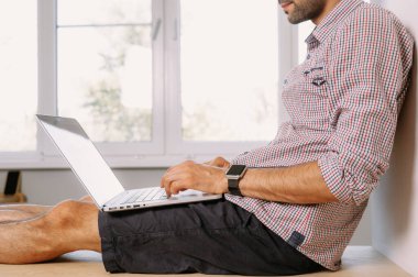 Gömlekli genç bir adam pencere eşiğinde oturuyor ve evde dizüstü bilgisayarla çalışıyor. Evde dizüstü bilgisayarla çalışan genç bir adam.