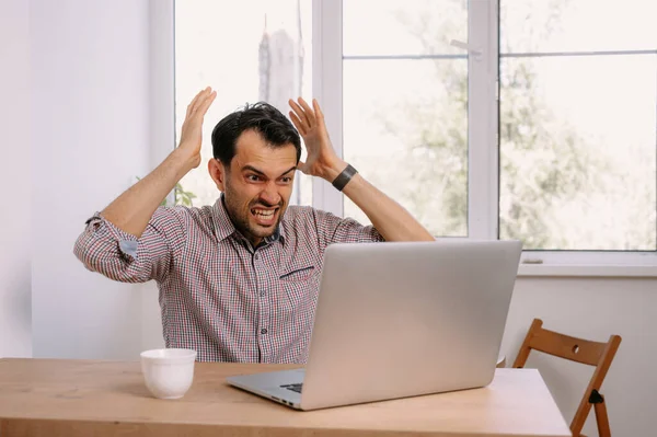 Angry man in shirt with a cup of coffee works on laptop.Young man working on laptop at home at the table.Young man working on his notebook.Working on project with laptop