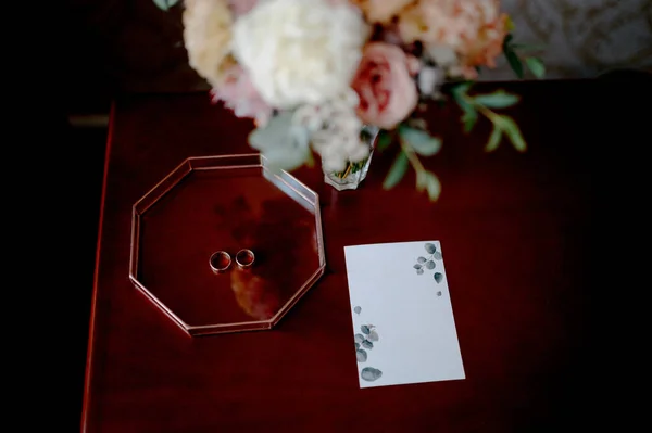 Due fedi nuziali d'oro, un mazzo di fiori e inviti nuziali su un tavolo di legno — Foto Stock