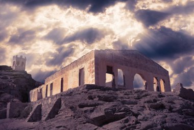 terk edilmiş eski taş ev ve dramatik bir bulutlu gökyüzü altında bir kaya üzerinde küçük bir kule