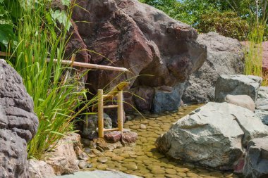 Bamboo sound water fountain next to calm languid stream surrounded natural stones as part of traditional Japanese Zen Garden. Traditional upright natural bamboo water spout . clipart