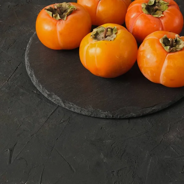 Group of fresh orange ripe persimmon fruits laying on round black slate on a dark background texture and copy space. — Stock Image Group of fresh orange ripe persimmon fruits laying on round black slate on a dark background texture and copy space. Stock Image