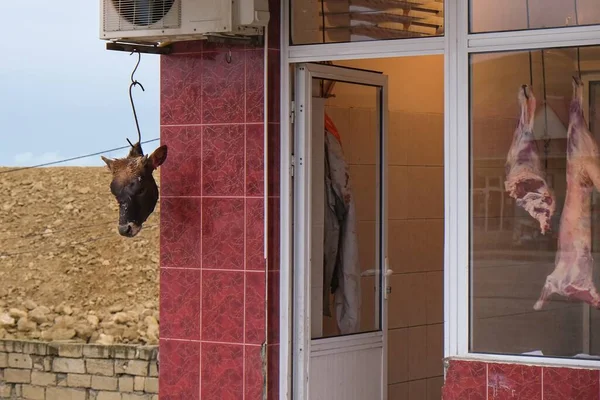Θέα μέσα από βρώμικο θολή τζάμι παράθυρο στο νωπό κρέας σφάγια κρέμονται και κομμένο κεφάλι σε γάντζο στο δρόμο κρεοπωλεία κατάστημα στην Caucasus χωριό αγορά. — Φωτογραφία Αρχείου