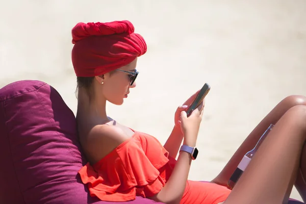 サングラス ターバンで美しいセクシーな女性がビーンバッグの砂の上に座って スマートフォンにメッセージを書き込みます — ストック写真
