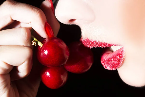 Sexy woman eating cherry. Sensual red lips in sugar isolated on black background.
