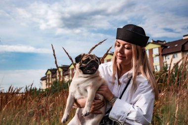 Genç ve mutlu bir kadın şehir parkında koşum takımı ve siyah bir tasmayla sevgili köpeğini kucaklıyor. Sokak fotoğrafçılığı. En sevdiği evcil hayvanlar. Gökyüzü arkaplanı.