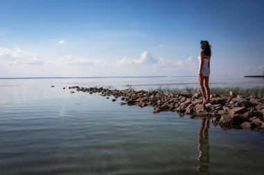 Genç, seksi, güzel bir kadın sahildeki kayalıklarda duruyor ve dinleniyor, sıcak güneşin tadını çıkarıyor ve suyun üzerinde günbatımının tadını çıkarıyor. Turizm, tatil. Dikiz aynası. Arkaplan.