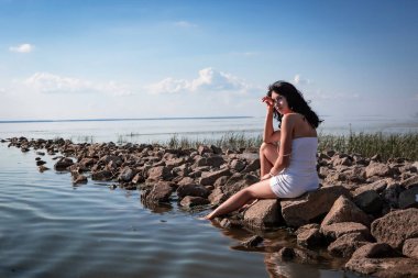 Genç, seksi, güzel bir kadın sahildeki kayalıklarda oturuyor ve dinleniyor, sıcak güneşin tadını çıkarıyor ve suyun üzerinde günbatımının tadını çıkarıyor. Sağlık, turizm, tatil, hafta sonu. Arkaplan