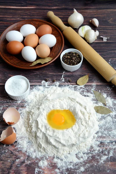 Egg in flour, rolling pin, eggs on a plate, bay leaf, salt and pepper on a dark wooden background, top view.