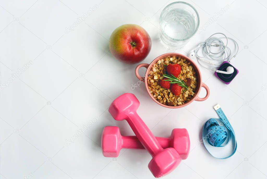 Dumbbells, measuring tape, glass of water, player and headphones for running, bowl of muesli and an apple on a white background, top view.