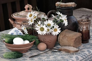Koyu arka planda bir masa örtüsü ile kaplı bir masada bir köy hala yaşam şeklinde bir kil sürahi, ekmek, yumurta, salatalık papatya çiçekleri.