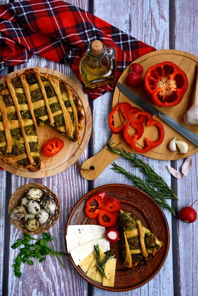 Appetizing vegetable pie, brown plate with a piece of pie, cheese and pepper, chopping board with sliced pepper, garlic, a bottle of olive oil, quail eggs and greens on a wooden table, top view.