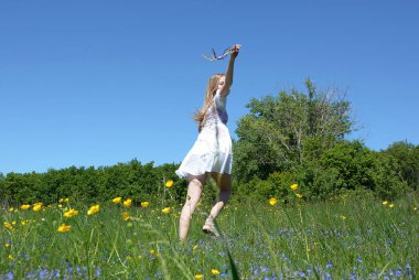 Akan sarı saçlı bir kız çiçekli bir çayır ile çalışır ve elinde çok renkli kurdeleler tutar, arka görünümü.