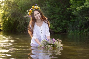 Akan koyu saçlı ve kafasına çelenk ile güzel bir genç gülümseyen kız bir nehirde duruyor ve su üzerinde çiçek çelenk tutuyor.