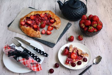 Ev yapımı yaz çay partisi konsepti. Çilekli turta, çaydanlık, sofra takımı, tabaklar ve hafif ahşap arka planda taze böğürtlenli kase. Üst görünüm