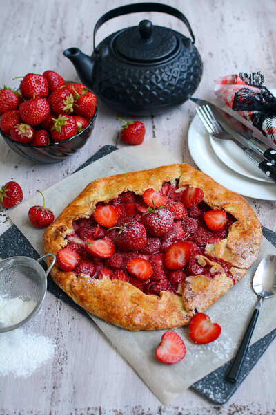 Ароматный торт со свежей клубникой, тарелками, ложками, чайником на деревенском фоне. По вертикали. Летняя выпечка и домашний чай. Вид сверху
