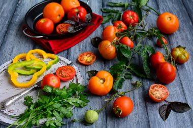 Dalları olan sulu olgun domatesler, sebzeli metal bir kase ve gri kırsal bir masada maydanoz ve bıçaklı bir kesme tahtası. Sebze pişirme süreci. Kapat.
