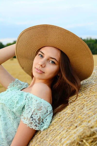 Uma menina de aparência europeia em um chapéu e uma blusa turquesa fica no fundo de pilhas de palha e sorri para a câmera. Vertical — Fotografia de Stock