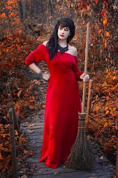 Portrait of a dark-haired woman in a red dress with a broom in her hands on a wooden bridge in a fairy forest. Vertical