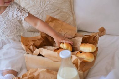 A little girl wants to take a baguette from a wooden box. Nearby is a bottle of milk. The girl is wearing a white lace dress. She is sitting in a bright room next to a lace pillow.