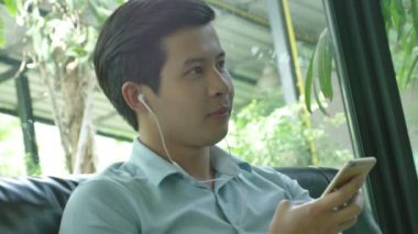 4k: Asyalı Genç adam kanepede rahatlatıcı ve smartphone ile müzik dinlemek