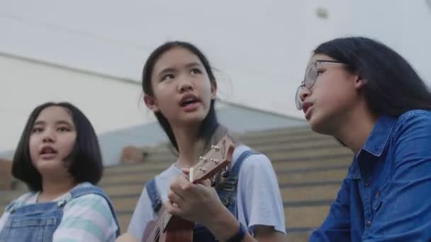 幸せなアジアの女の子の友人は ウクレレを演奏し 学校で一緒に歌を歌う楽しみを持って 60Fps — ストック動画