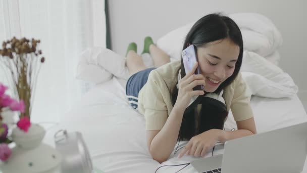 肖像可爱的亚洲女孩躺在床上 并使用笔记本电脑和手机沟通 — 图库视频影像