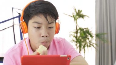 Tablet bilgisayarınızla turuncu kulaklık, elma parça yemek ve tablete izlerken Teenage çocuk oynamaya mutlu Asyalı çocuk.