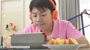Tablet bilgisayarınızla turuncu kulaklık, elma parça yemek ve tablete izlerken Teenage çocuk oynamaya mutlu Asyalı çocuk.