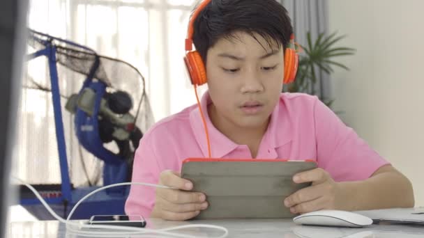 快乐的亚洲男孩玩平板电脑与橙色耳机 十几岁的男孩吃一块苹果和观看平板电脑 — 图库视频影像