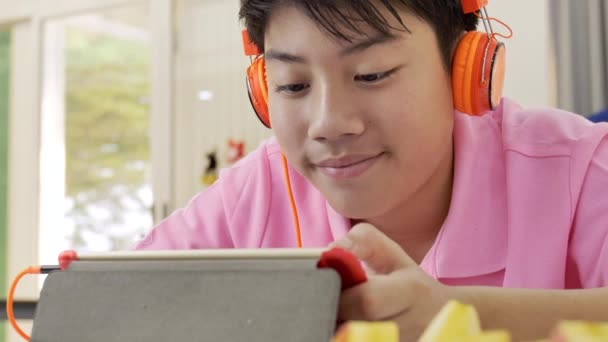 幸せなアジアの少年少年リンゴの部分を食べると タブレットで見ているオレンジのヘッドフォンでタブレット コンピューターで再生 — ストック動画