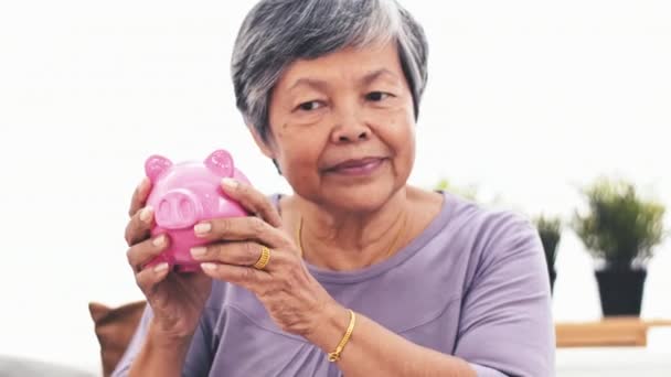 陽気なアジアの年配の女性はカメラに笑顔の貯金を揺れします 節約貯金箱屋内を保持している大人の女性の肖像画 — ストック動画