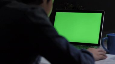 Masabaşında dizüstü bilgisayar kullanan genç bir çocuğun omzundan çekilmiş fotoğrafı, yeşil ekrana bakıyor. Dolly atış 60fps.