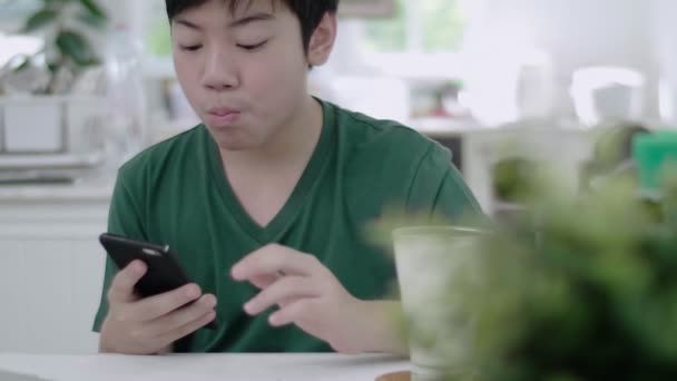 スマートフォンのアプリケーションを通じてスクロールを使用するアジアの若い男の子は ゆっくりと自宅でパンビスケットとチョコレートでオーガニックミルクの朝食を食べます — ストック動画