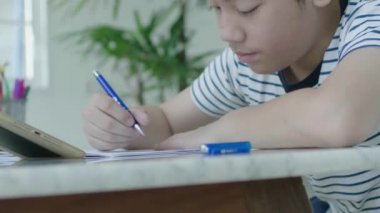 Asyalı çocuk cevapları bulmak için bir tablet kullanarak ev ödevi yapıyor evde bir sandalyeye oturur.