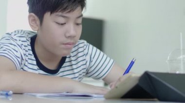 Asyalı çocuk cevapları bulmak için bir tablet kullanarak ev ödevi yapıyor evde bir sandalyeye oturur.