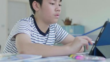 İyi görünümlü Asyalı çocuk evde bir sandalyeye otur ve cevapları bulmak için bir tablet kullanarak ev ödevini yapmak.
