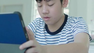 İyi görünümlü Asyalı çocuk evde bir sandalyeye otur ve cevapları bulmak için bir tablet kullanarak ev ödevini yapmak.